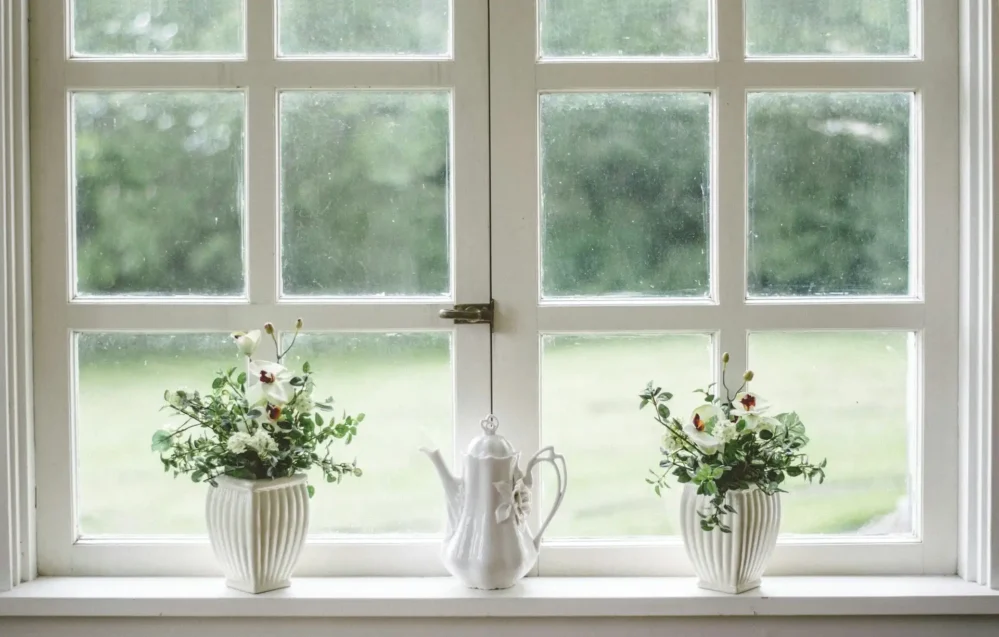 White-paned window with two vases of flowers and a decorative white teapot on the sill, overlooking a green outdoor view